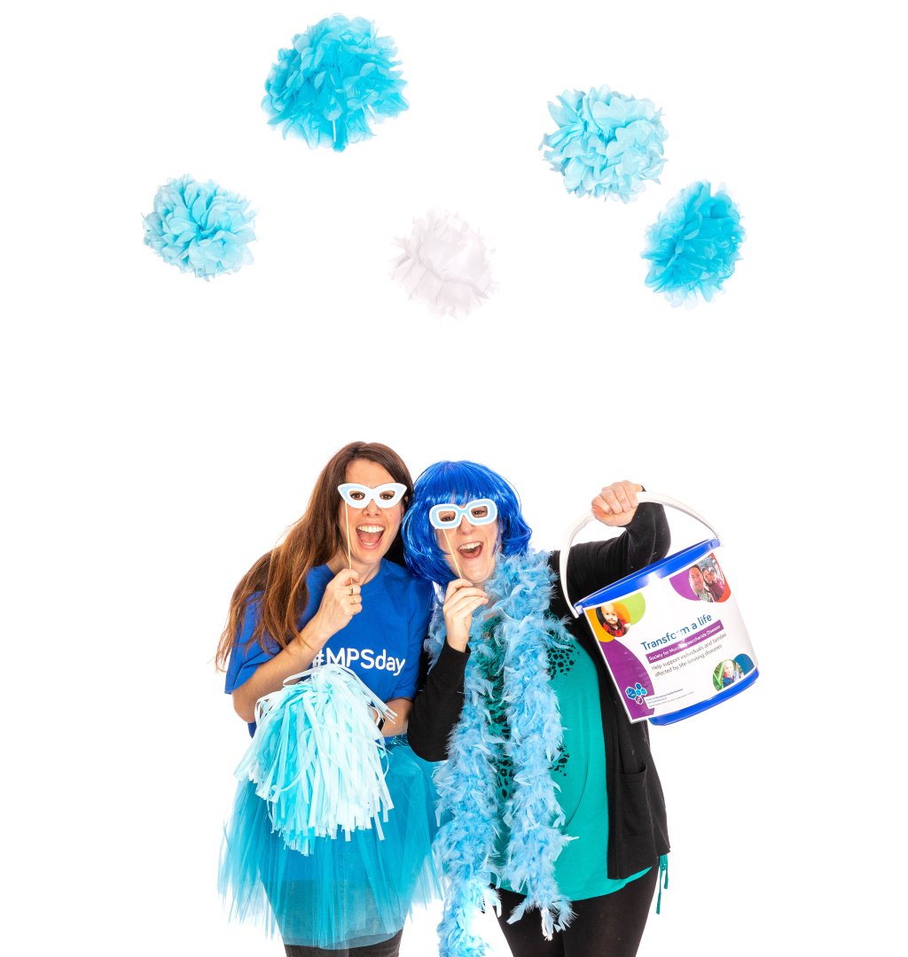 Two women wearing blue cardboard masks with blue t-shirts, a blue wig, pom-poms and a feather boa hold a fundraising bucket for MPS Society.
